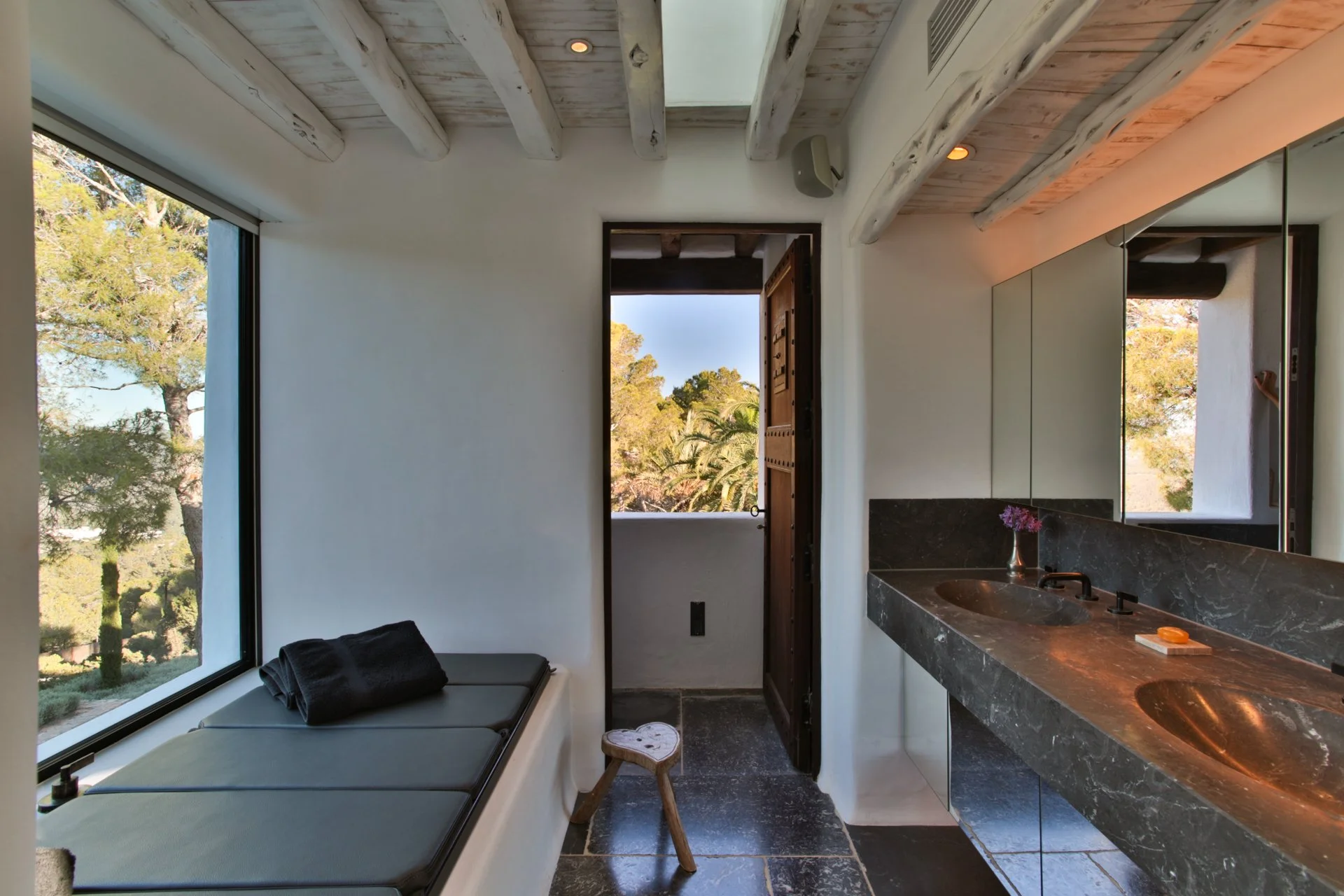 Interior view of a bathroom with a black marble countertop, seeing a sink with a faucet, and a mirror on the wall. There is a window showing trees outside, and a door leading to a balcony or outdoor area. A cushioned window seat with a folded black t
