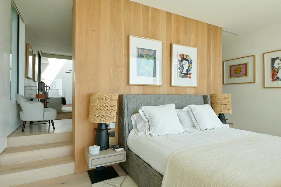 Bedroom with a gray upholstered bed, white bedspread, and pillows, flanked by two nightstands with matching lamps and art on the wall, including a wooden accent wall and a sitting area with armchairs visible through a doorway.