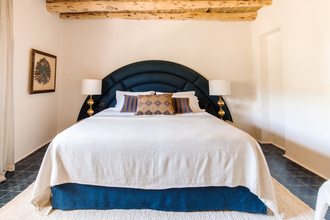 A neatly made bed with a dark blue upholstered headboard, white bedding, and decorative pillows with traditional patterns. Two matching bedside lamps with white shades sit on gold bases. The room has white walls, a framed artwork on the left, and a n