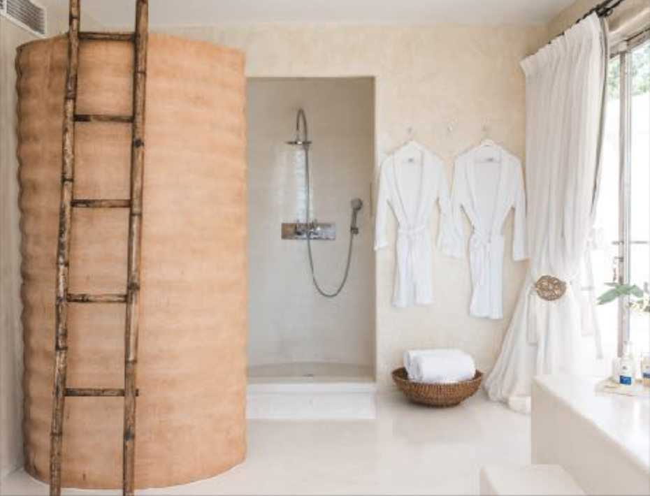A bathroom with a curved shower, two white robes hanging on the wall, a basket with towels, and a window with white curtains.