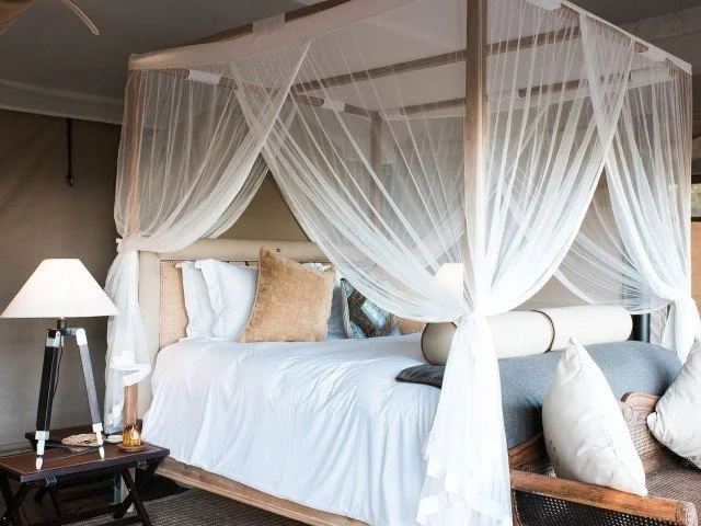 A four-poster bed with white sheer curtains, white bedding, and multiple pillows in a bedroom with a bedside table and lamp.