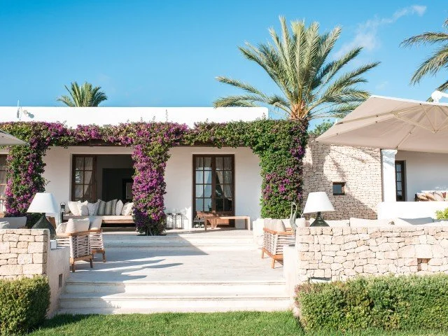 Exterior view of a villa with white walls, brown doors, purple bougainvillea, palm trees, and outdoor seating under umbrellas on a sunny day.