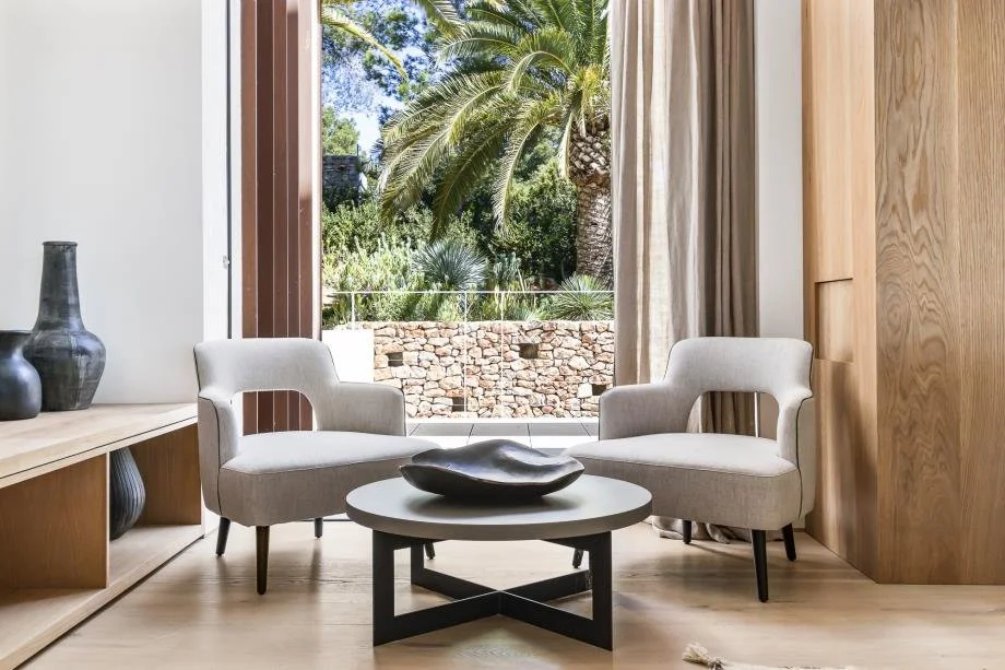 Living room with two beige armchairs, a round coffee table with a black decorative bowl, wooden cabinets, a yellow curtain, and a large window showing outdoor trees and a stone wall.