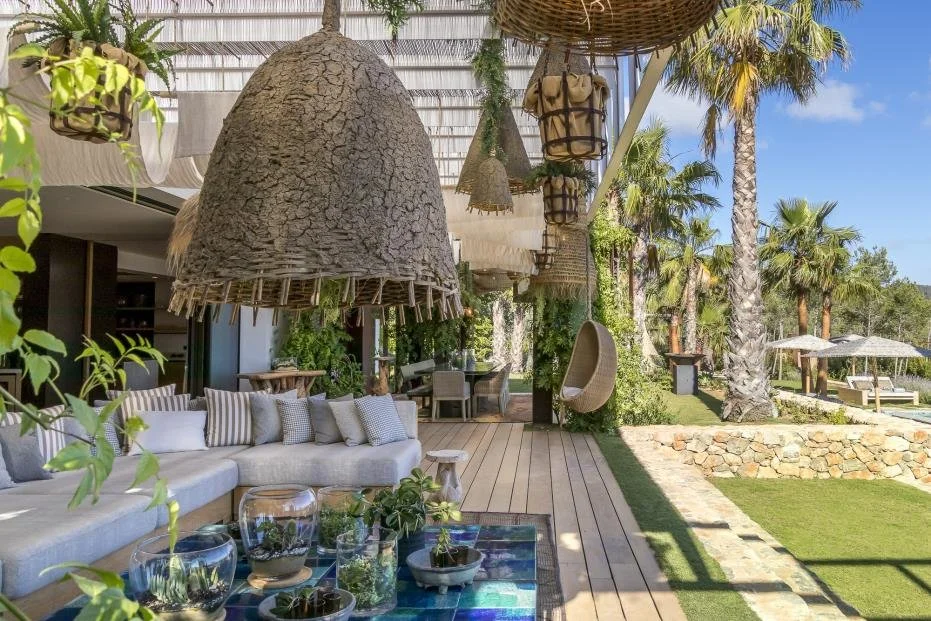 Outdoor patio with beige sectional sofa, hanging wicker lamps, glass bowls with plants, wooden deck, lush palm trees, and a grassy area with lounge chairs and umbrellas