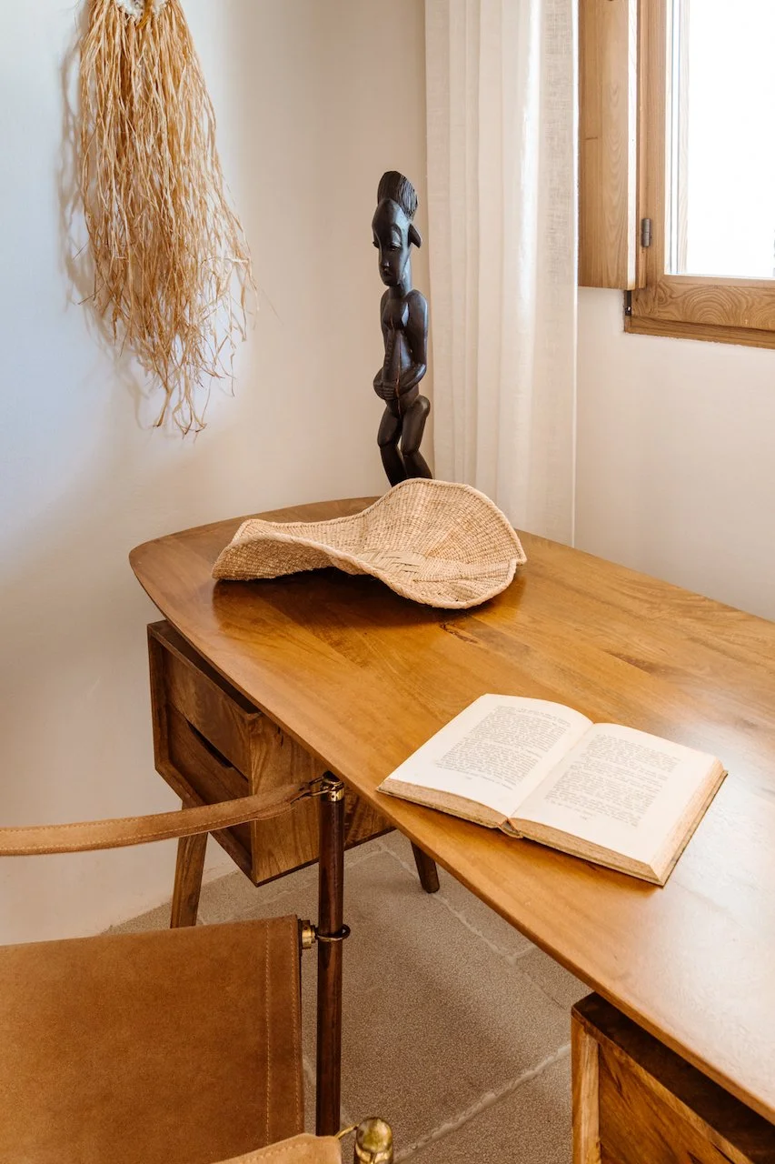 A wooden desk with an open book, a straw hat, and a carved wooden sculpture of a person, situated near a window with a curtain and a wall hanging made of natural fibers.