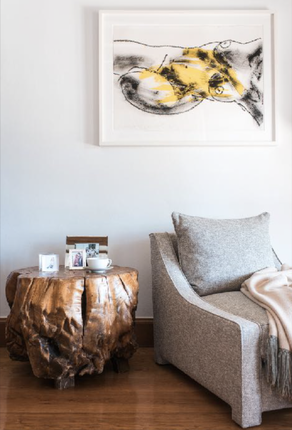 A living room corner with a framed abstract art piece featuring black and yellow brushstrokes above a gray upholstered armchair. A rustic wooden side table holds framed photographs and a white teacup with a saucer.