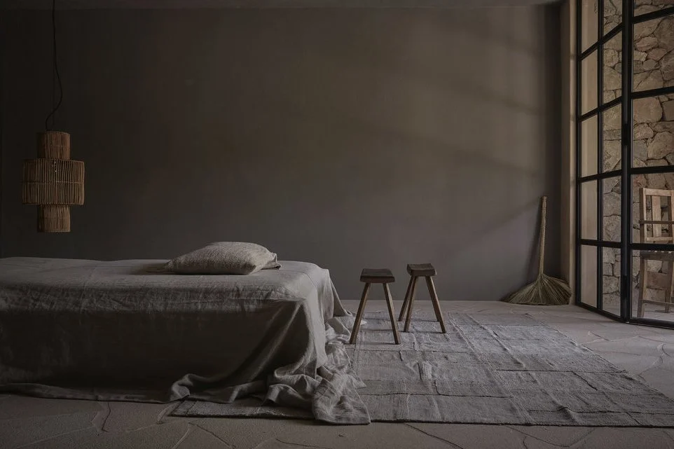 A minimalistic bedroom featuring a bed with beige linens, two small wooden stools, a textured rug, and a broom in the corner. The room has large glass doors with black frames, and a stone wall is visible outside.
