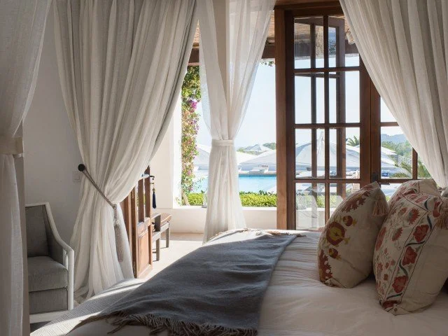 A bedroom with beige curtains, a bed with decorative pillows, and a view of boats and water outside through a wooden window door.
