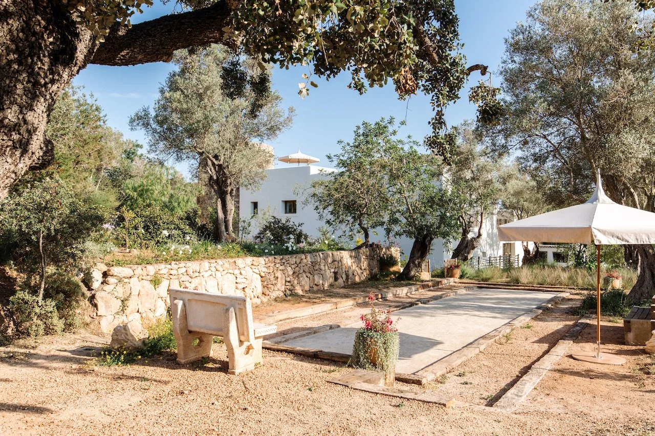 A peaceful outdoor scene with a bocce ball court, a stone bench, and large umbrellas for shade, surrounded by trees, plants, and a white building in the background.