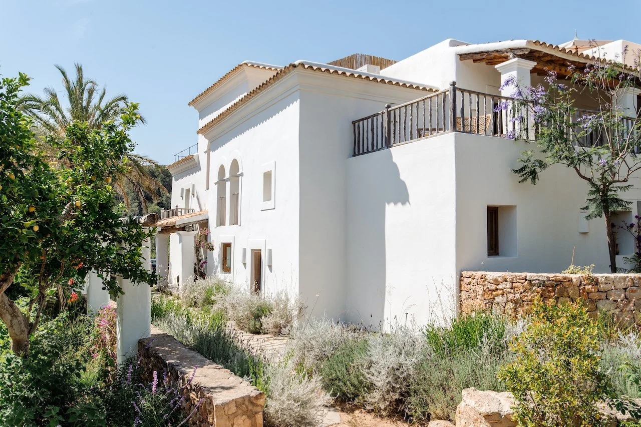White Mediterranean-style house with terraces and wooden balcony railings, surrounded by green plants, trees, and a stone wall under a clear blue sky.
