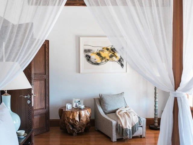 A cozy corner with a gray armchair and a throw blanket, a wooden side table with candles and a photo, a lamp, and a white sheer curtain tied aside revealing a white wall with abstract art.