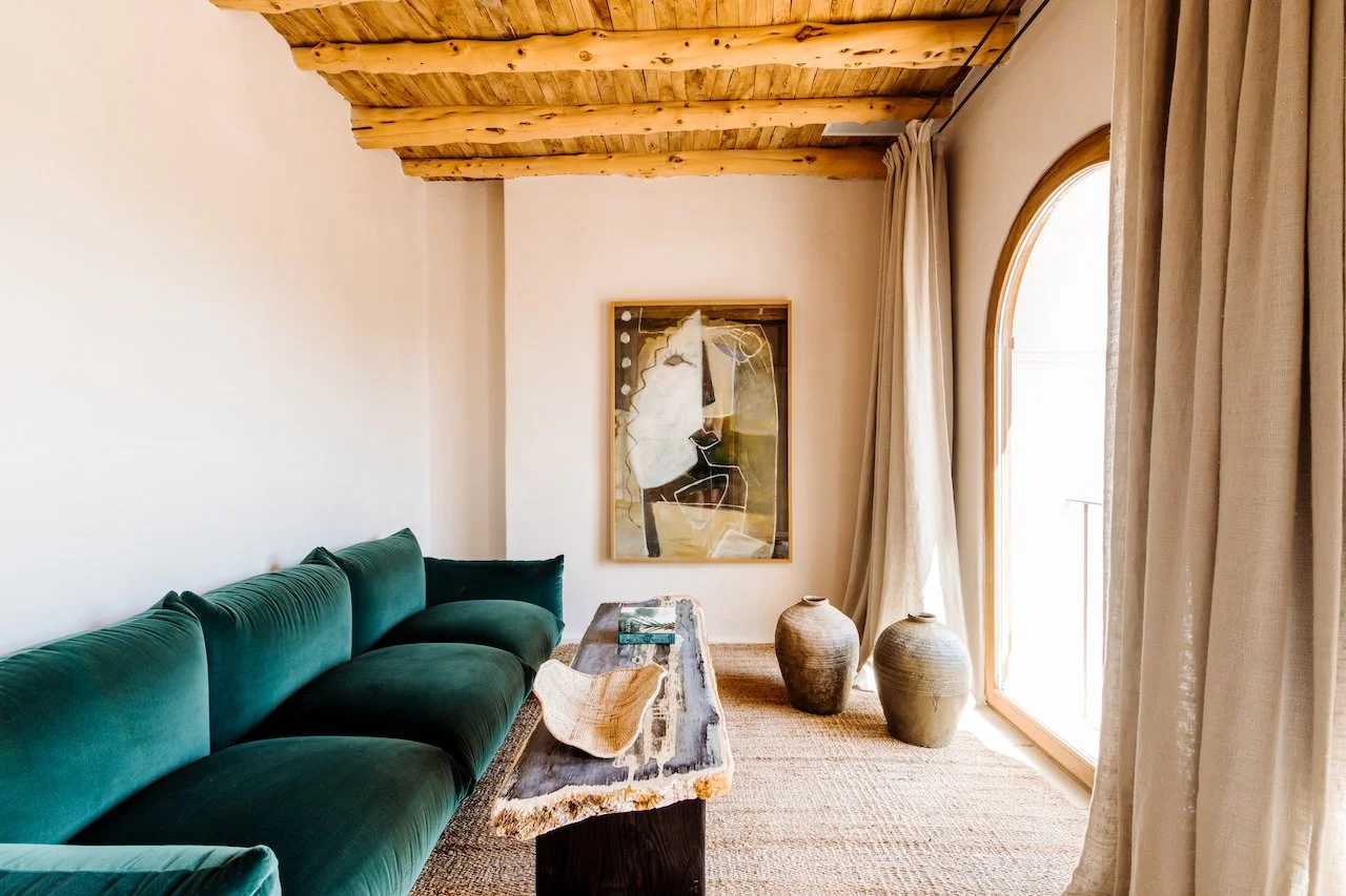 Living room with a green velvet sofa, a rustic wooden coffee table, abstract art on the wall, two large ceramic pots near a large window with beige curtains, and a wooden ceiling.