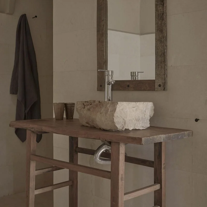 Rustic bathroom sink with a large stone basin mounted on a wooden vanity, with a mirror above it, and towels hanging on hooks on the wall.