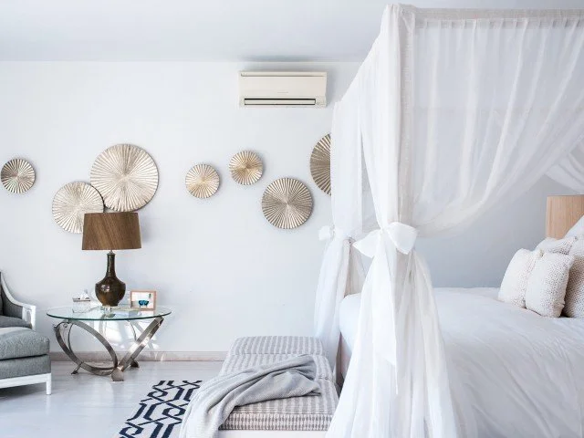 A bedroom with white walls and bedding, a four-poster bed with white curtains, a gray bench with a throw blanket, wall decor with round metallic wall art, a side table with a lamp and books, and an air conditioning unit on the wall.