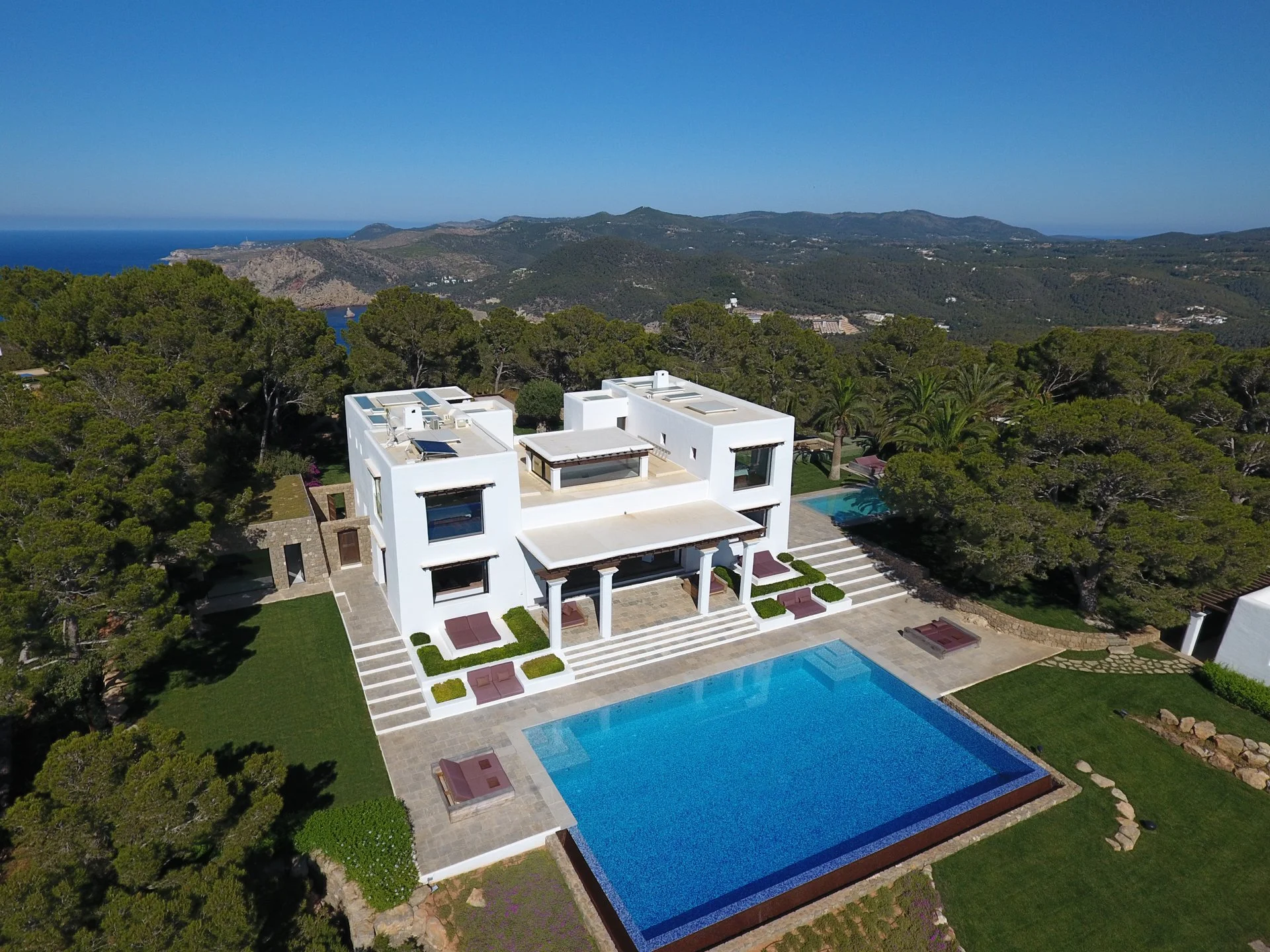 Aerial view of a modern white house with a large swimming pool, surrounded by greenery and hills, with ocean in the background under a clear blue sky.