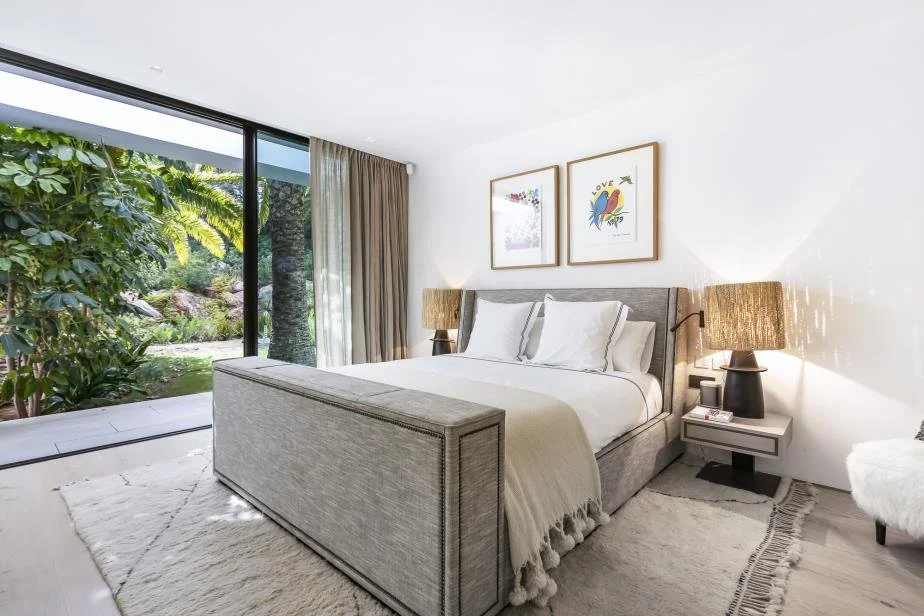 Modern bedroom with a large bed, two bedside tables with lamps, framed artwork above the bed, and a sliding glass door leading to a lush outdoor garden.