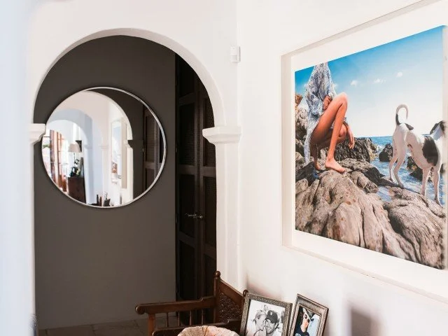 A white-walled room with a large beachside photograph on the wall showing a woman with a dog on rocks near the ocean. There is a round mirror on the dark foyer wall, a wooden chair with framed photos, and an open door to a closet.