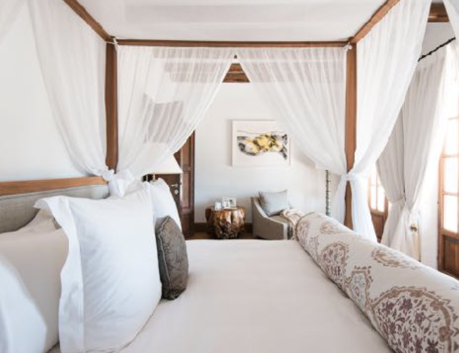 A cozy bedroom with a four-poster bed with white curtains, a stone side table, and a small sitting area near a window.