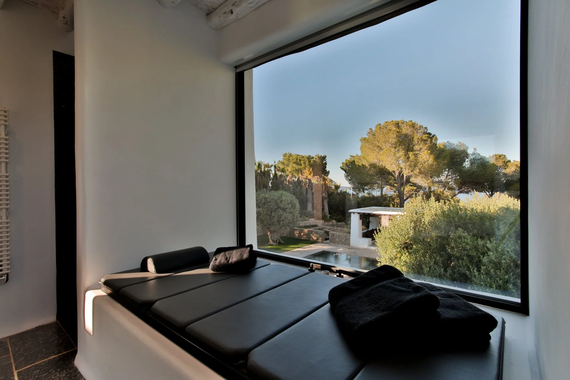 Interior view of a room with a large window overlooking a garden with trees, bushes, and a small structure, featuring a black cushioned bench under the window and towels on top.