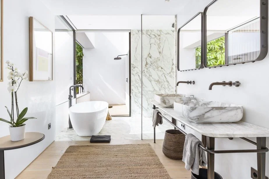 Modern bathroom with white walls, marble countertops, double vessel sinks, and a freestanding bathtub, with large mirrors and a glass shower enclosure.