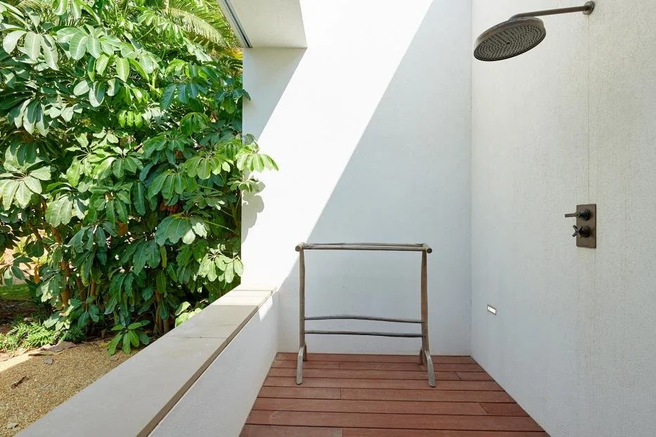 Outdoor shower with a rain shower head, a control panel, and a wooden floor, surrounded by beige walls and lush green trees.