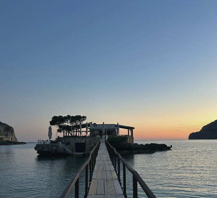 A wooden pier leading to a seaside restaurant during sunset, with trees and umbrellas on the terrace.
