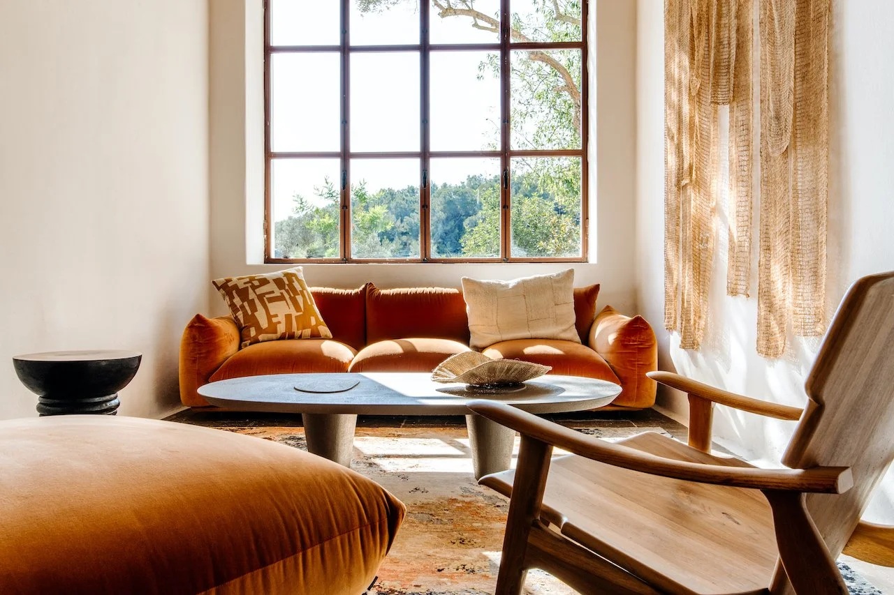 Living room with a large window, orange velvet sofa with pillows, wooden armchair, wooden coffee table with decor, and sheer curtains, sunlight streaming in.