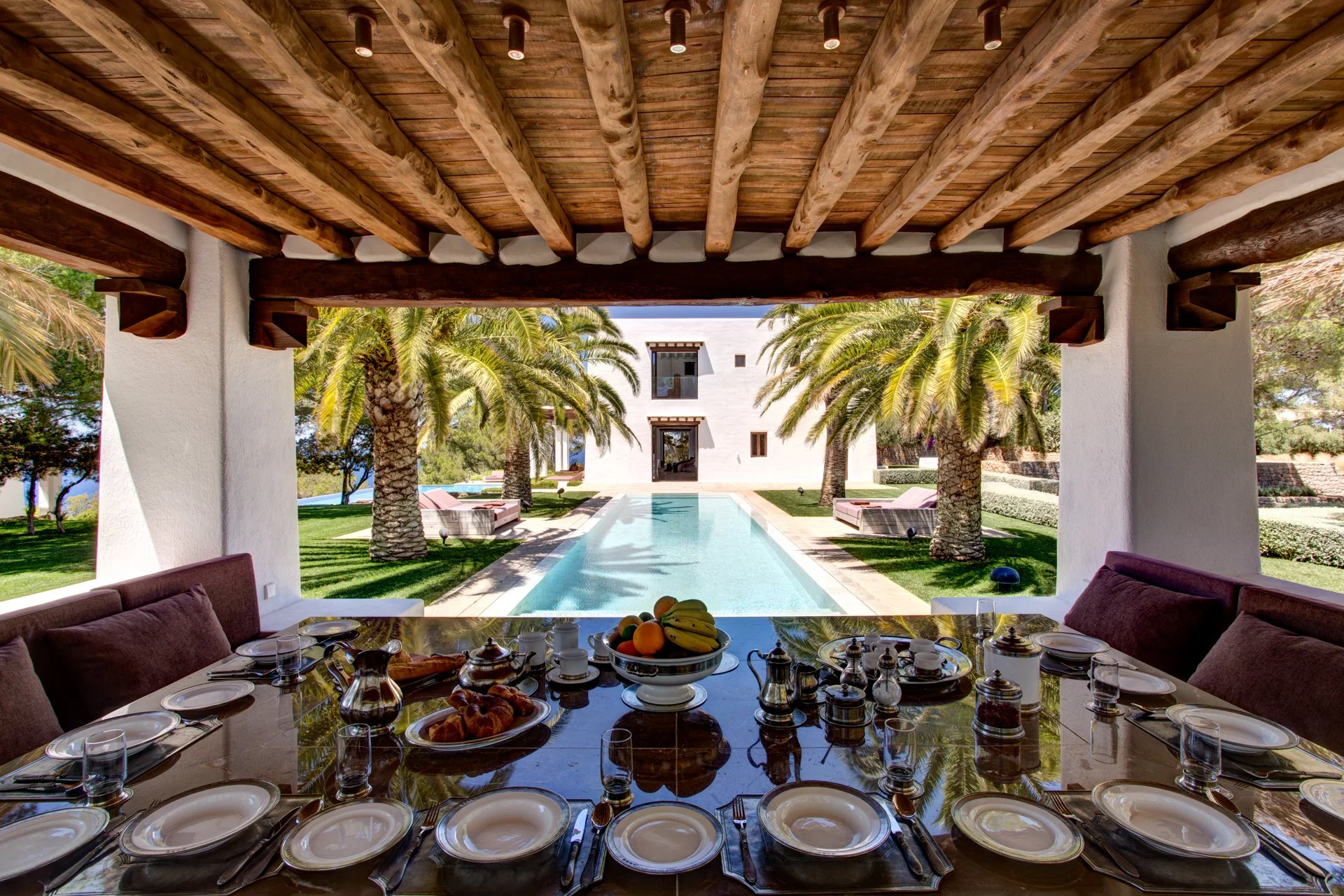 Dining table set with plates, glasses, and food, overlooking a swimming pool flanked by palm trees in a backyard.