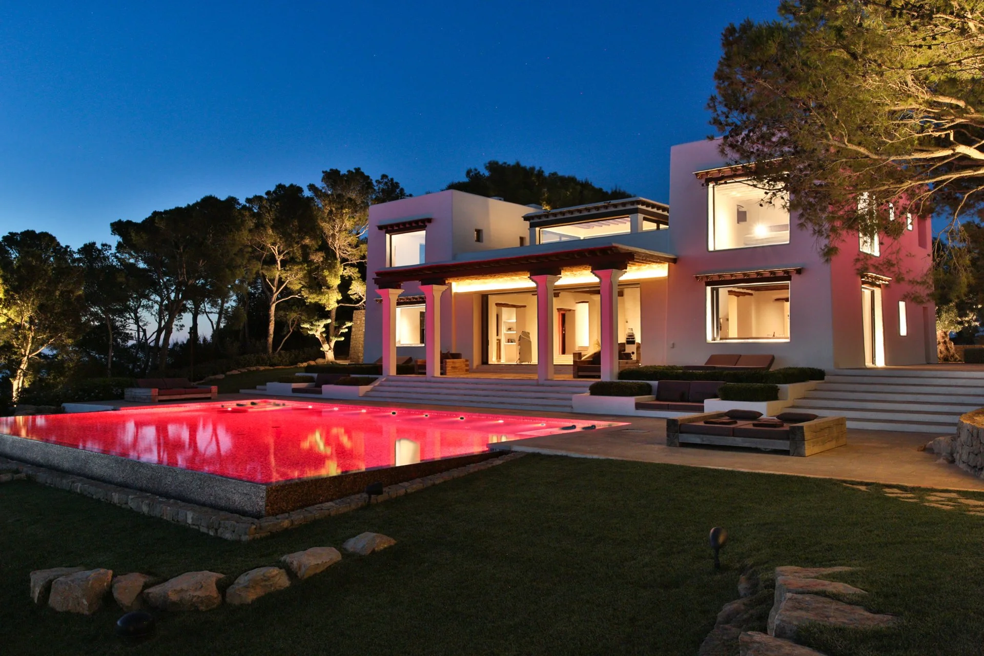 A modern two-story house at night with large illuminated windows, a lit porch, and a backyard with a swimming pool that has red lighting, surrounded by trees and rocks.