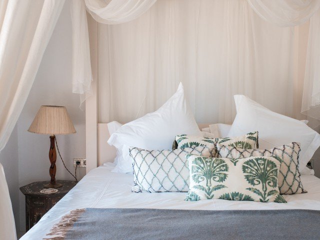 A neatly made bed with white pillows and decorative pillows with green nature-inspired patterns in a cozy bedroom with a soft lighting lamp on a bedside table.