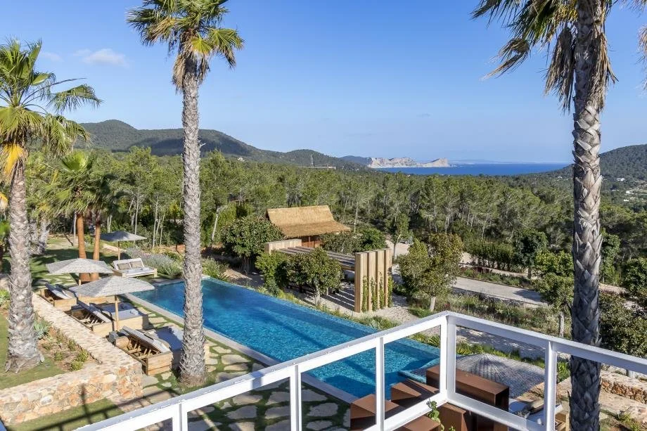 View of a luxurious outdoor swimming pool surrounded by lounge chairs with umbrellas, palm trees, lush greenery, hills, and a distant ocean in the background.