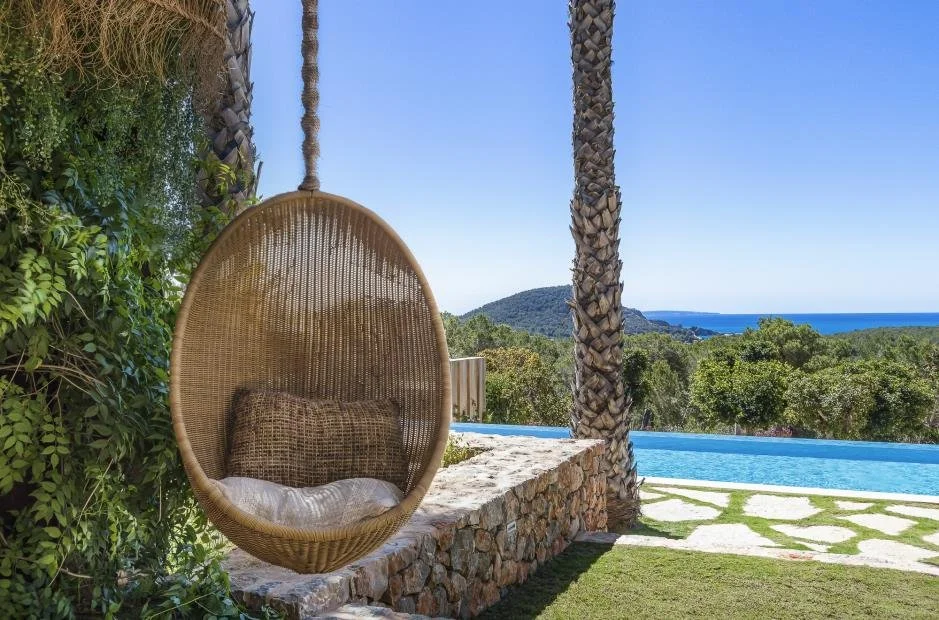 A hanging wicker swing chair with cushions, next to a stone wall, overlooking a pool, with a view of trees, hills, and the ocean in the distance under a clear blue sky.