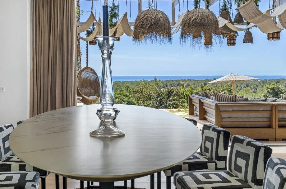 Dining area with a view of greenery and ocean in the background, decorated with hanging lamps and a transparent candle holder on the table.
