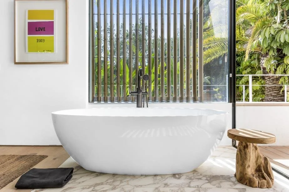 Modern bathroom with a white freestanding bathtub, a wooden stool, a framed colorful art piece, and large windows showing lush greenery outside.