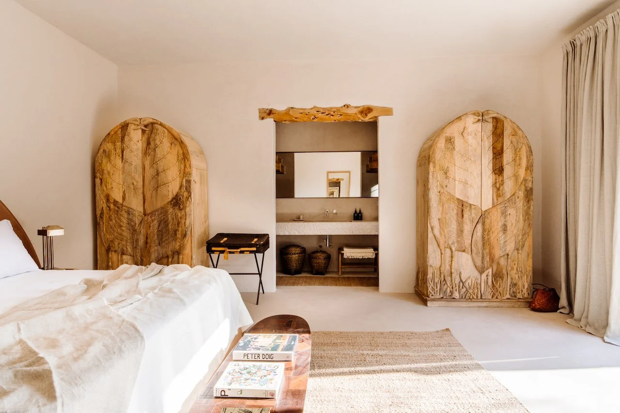 A minimalist bedroom with a bed, two large wooden carved panels with abstract designs, a bedside table holding books, and a bathroom with a vaulted mirror and baskets visible through an open doorway.