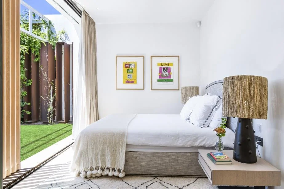 A bright bedroom featuring a bed with white linens, two framed colorful art pieces on the wall, a bedside table with a flower vase and books, a large window with curtains, and a view of a small outdoor yard with green grass and a trellis.