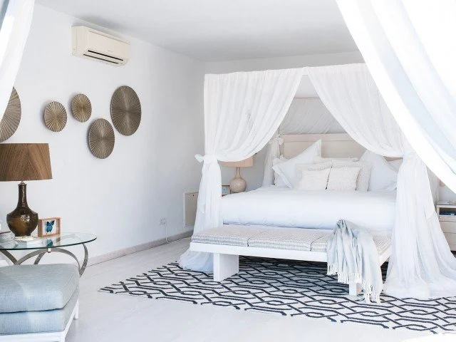 A white bedroom with a four-poster bed, white curtains, a bedside lamp, wall decor, a glass table, and a patterned rug.