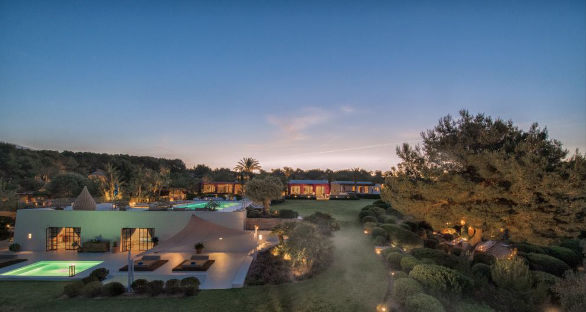 Luxury resort at dusk with illuminated swimming pool, palm trees, and modern buildings, surrounded by lush greenery and trees.