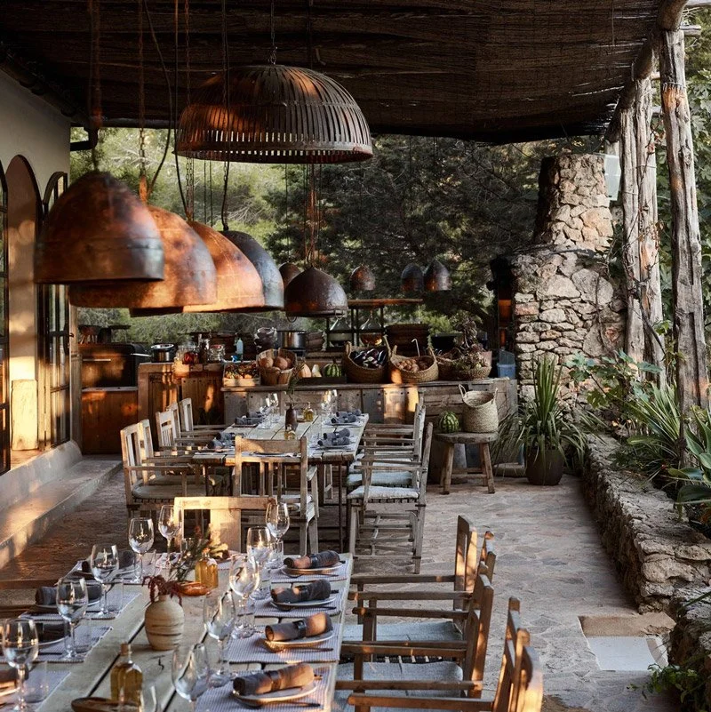 Restaurant with rustic decor, large hanging copper lamps, wooden tables set with wine glasses and napkins, surrounded by greenery and stone accents.