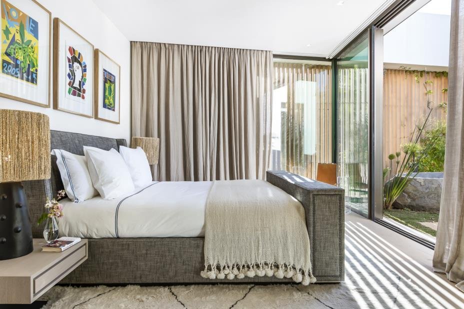 Modern bedroom with a large bed, white bedding, and decorative pillows, next to a sliding glass door leading to a patio with outdoor plants.