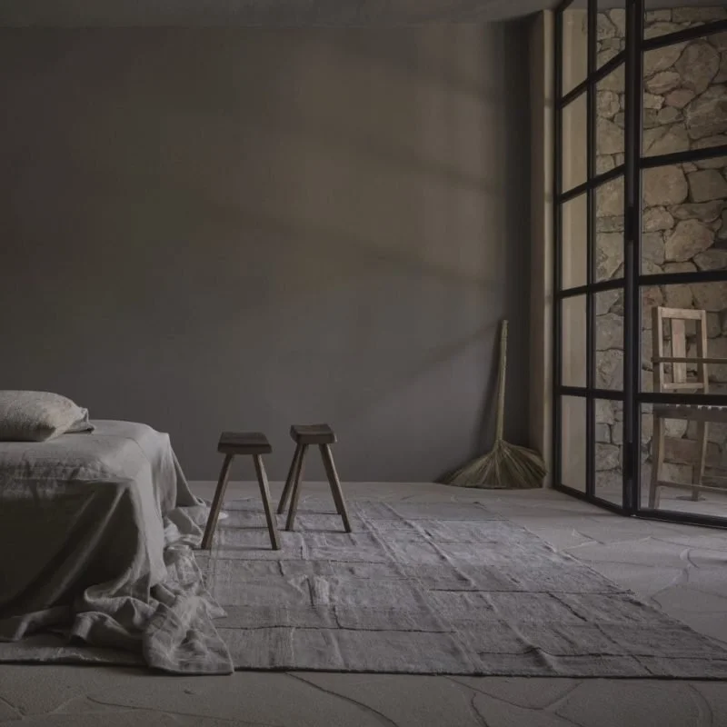 Minimalist bedroom with a bed on the left, two small stools in the center, a broom in the corner, and a large glass door leading to a stone wall patio.