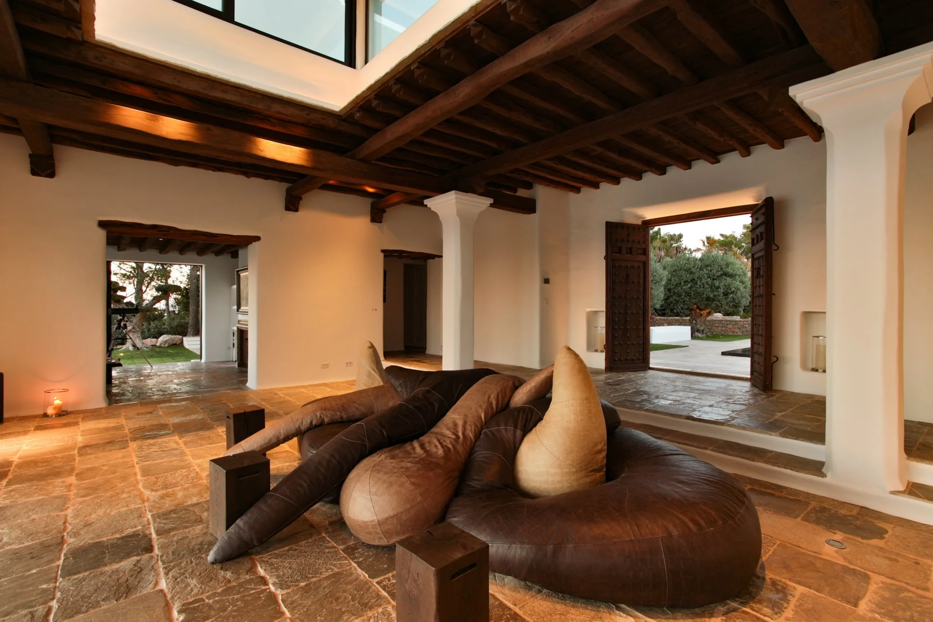 Living room with exposed wooden beams, large leather sectional sofa with beige pillows, open wooden doors leading outside to a garden with trees, tiled floor, and a large window letting in natural light.