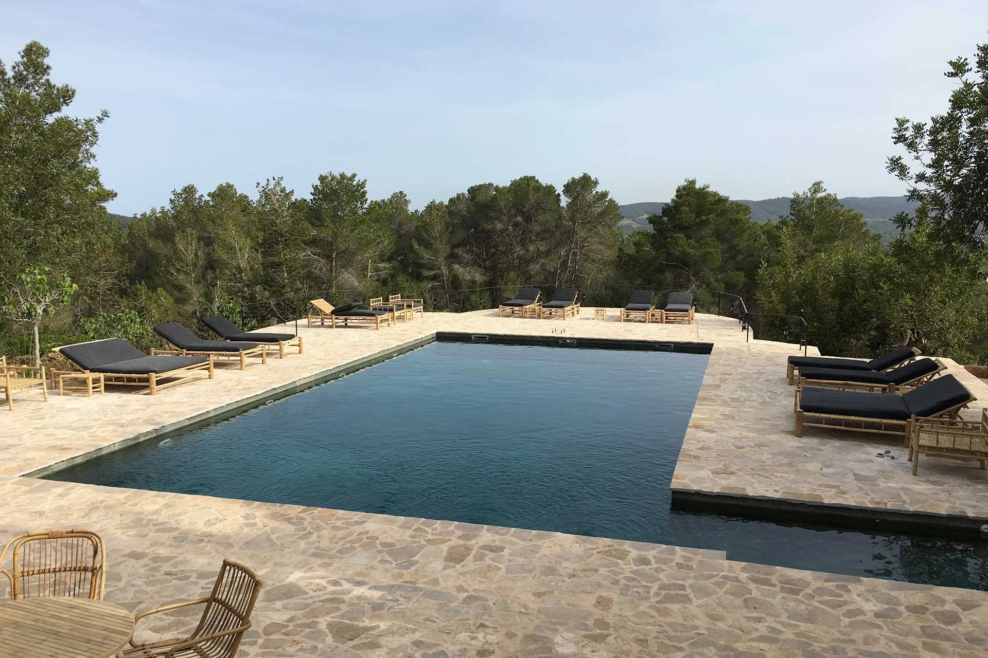 A swimming pool outdoor area with lounge chairs on a stone patio, surrounded by trees and hills in the background on a clear day.