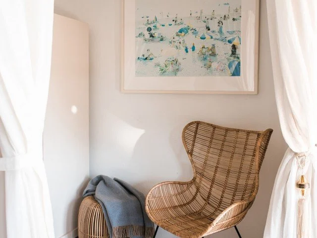 A wicker chair with a gray throw blanket, positioned near white curtains and a framed artwork of a beach scene on a white wall.