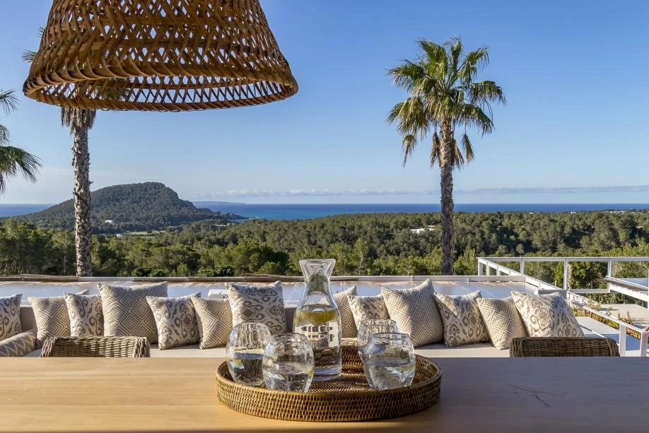 A patio with a long sofa filled with patterned cushions, a wooden table with a tray holding a glass pitcher and three glasses of water, a large wicker hanging lamp, two tall palm trees, lush green trees, a mountain, and a body of water in the distanc