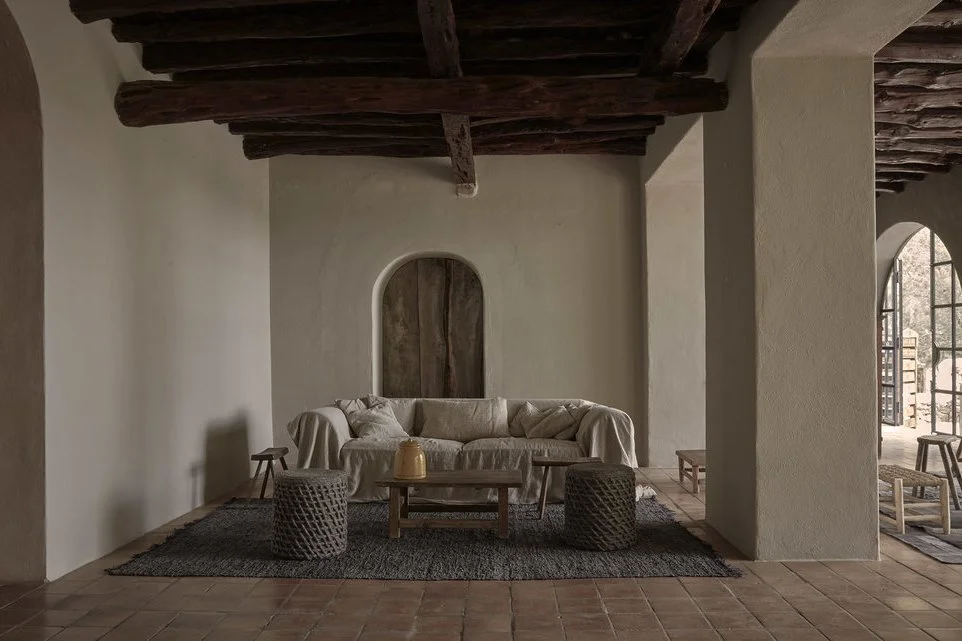 Living room with a beige sofa, two poufs, a wooden coffee table, and a dark rug, featuring wooden ceiling beams and arched doorways.