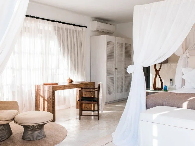 Bright hotel room with white curtains, a wooden desk, a chair, a wardrobe, and a bed with white linens and a canopy.