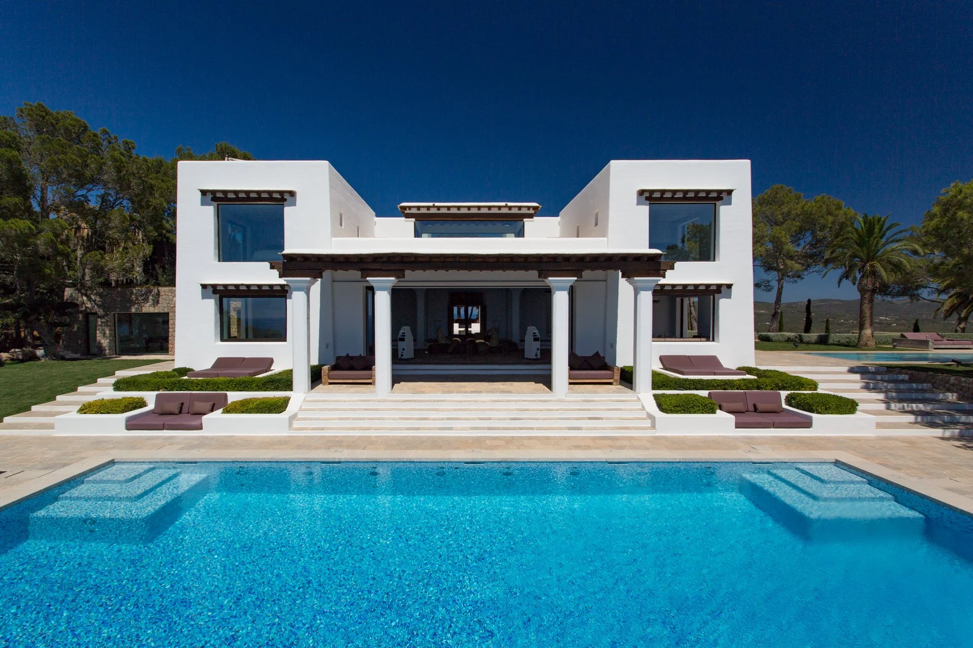 A modern white two-story house with a swimming pool in the foreground, surrounded by a landscaped garden with trees and outdoor seating, under a clear blue sky.