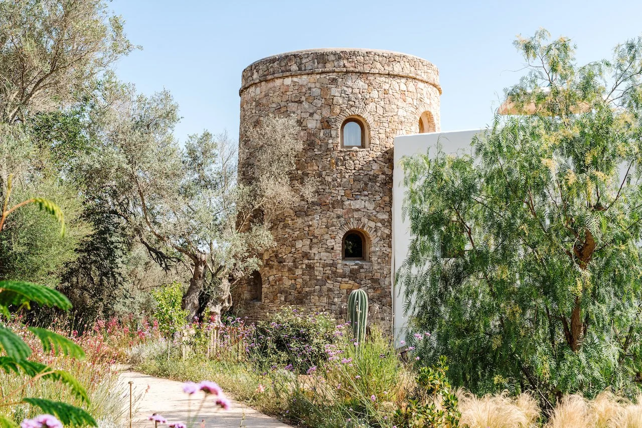 A stone tower with small arched windows in a garden with trees, flowers, and desert plants like cacti