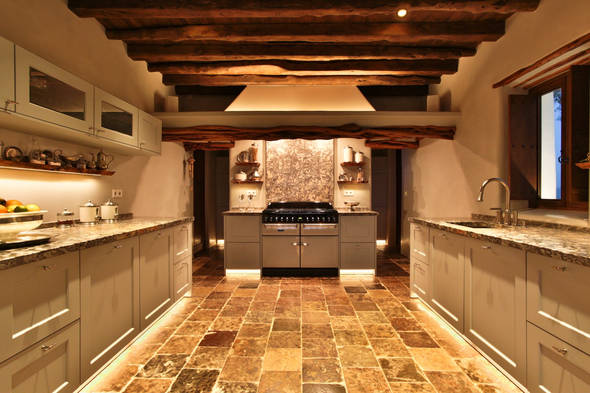 A rustic kitchen with wooden ceiling beams, white cabinets with glass doors, granite countertops, a black stove, and earth-toned tiled flooring; sunlight streams in from a window on the right.